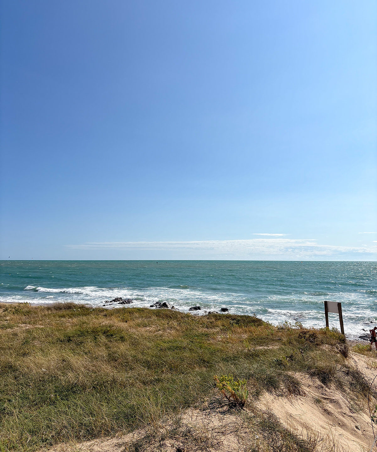 Strand mit Dünen und rauer See unter blauem Himmel.
